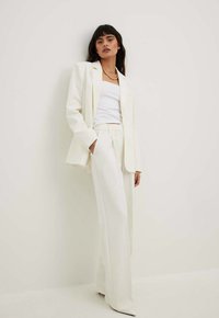 Tailored white suit with a long blazer and wide-leg trousers. Worn over a fitted white tank top. Smooth fabric with a crisp finish.