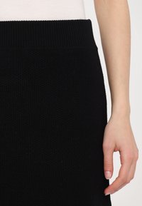 Gros plan d'une personne portant un haut blanc rentrée dans une jupe en tricot noire, avec un bras détendu visible sur le côté.
