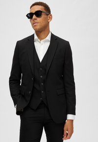 Schwarzer Dreiteiler mit slim fit, bestehend aus einem Jackett mit zwei Knöpfen, einem weißen Hemd und schwarzen Sonnenbrillen. Glattes Gewebe und maßgeschneiderter Schnitt.
