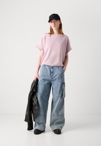 Světle růžový top s krátkým rukávem v kombinaci s modrými, oversized denimovými cargo kalhotami. Černá čepice a kožená bunda podtrhují ležérní vzhled.