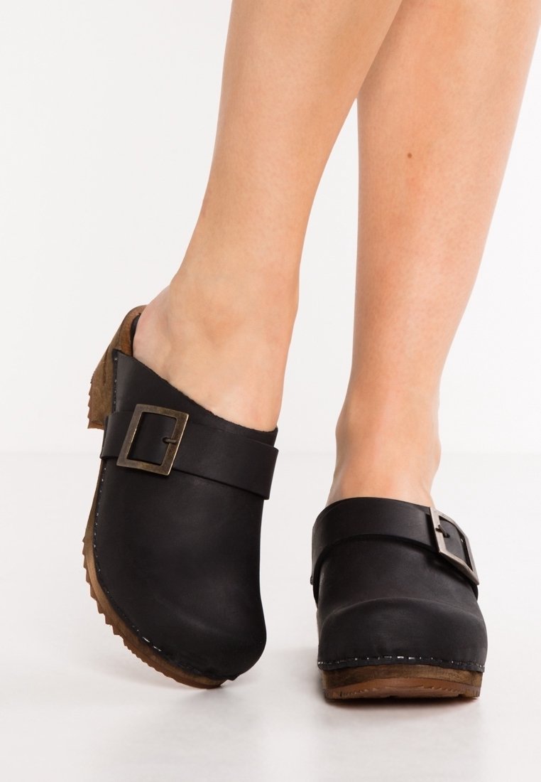 Jambes portant des sabots en cuir noir avec des boucles argentées et des semelles en liège marron sur fond blanc.