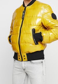 Bomber giallo lucido trapuntato con polsini e vita a coste nere, cerniera frontale e tasche laterali con zip, indossato sopra jeans grigio chiaro.