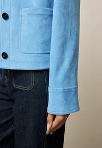 Veste en daim bleu clair avec poche plaquée à l'avant et boutons noirs, portée sur un jean en denim foncé, avec un gros plan sur la manche et la main.