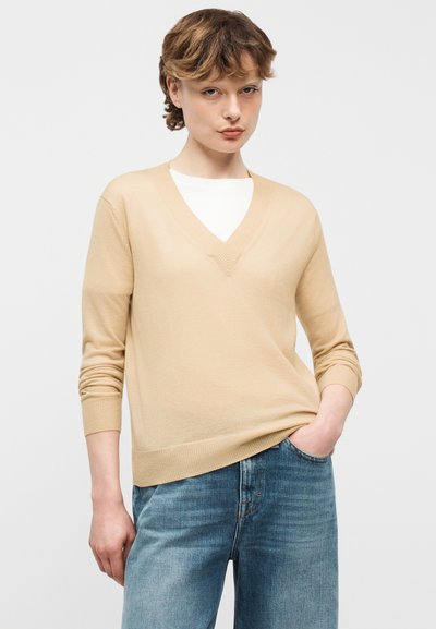 Jeune personne portant un pull beige à col en V par-dessus une chemise blanche et un jean bleu, avec une main dans la poche, sur un fond uni.