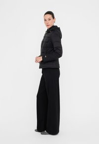 Giacca a vento nera con cappuccio, con zip frontale, design trapuntato e dettagli in metallo dorato, abbinata a pantaloni neri a gamba larga.