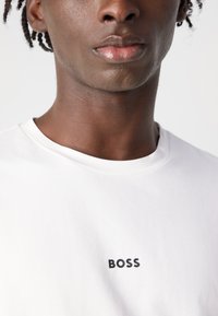 Camiseta de algodão branca com decote redondo, apresentando um logótipo preto "BOSS" impresso na frente. Textura suave, ajuste standard.