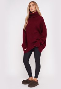 Burgundowa, oversizowa sweter z golfem, o teksturowanym splocie, w połączeniu z czarnymi legginsami i brązowymi butami wsuwanymi. Długie rękawy zakrywają dłonie.