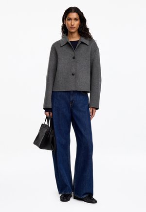 Femme portant une veste grise boutonnée, un jean large bleu foncé et des chaussures noires, tenant un petit sac à main noir dans sa main gauche.