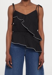 Schwarzes ärmelloses Oberteil mit weißen Rüschendetails und dünnen gebundenen Schulterträgern, getragen mit blauen hochgeschnittenen Jeans.