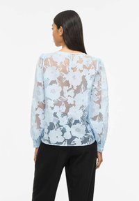 Femme portant un chemisier en dentelle florale bleu clair transparent avec des manches longues bouffantes, vue de dos, associé à un pantalon noir.