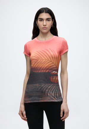 Femme aux longs cheveux foncés porte un t-shirt corail avec un coucher de soleil et des ombres de feuilles de palmier sur une eau sombre, associé à un pantalon noir.