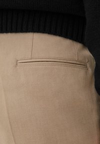 Beige bomullbyxor med en närbild av bakfickan som har en sydd horisontell linje, i kombination med en texturerad svart tröja.