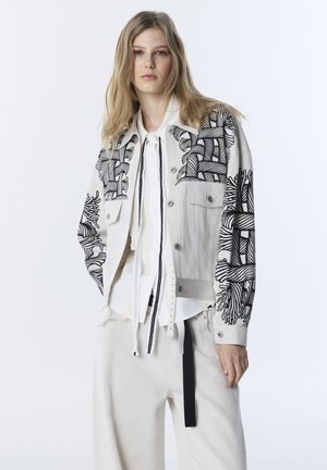Mujer con chaqueta beige clara con estampados botánicos abstractos en negro, camisa blanca con detalles de encaje y pantalones sueltos de color claro con trabilla negra.