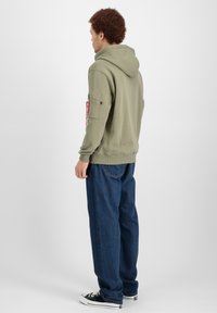 Felpa verde oliva con cappuccio e tasca con zip sulla manica, polsini a coste e vestibilità comoda, abbinata a jeans in denim blu a gamba larga.