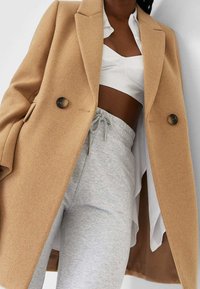 Wollmischung-Cape aus Kamelhaar mit Reverskragen und zwei großen Knöpfen, getragen über einem weißen Cropped-Top und grauen Sweatpants, was einen gestapelten Look zeigt.