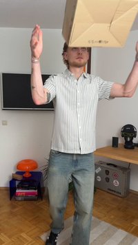 Person som balanserer en pappeske på hendene. Har på seg en hvit og grå stripete kortermet skjorte og lyseblå jeans. Moderne innendørs omgivelser.