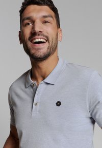 Lichtgrijs polo-shirt met een textuurfinish, voorzien van een twee-knops knoopsluiting en een hexagonaal zwart logo accent op de borst.
