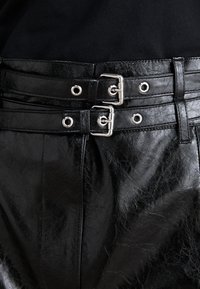 Pantaloni neri in finta pelle a vita alta con chiusura a doppia fibbia, caratterizzati da accessori in metallo argentato e una finitura testurizzata.