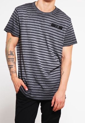 Mężczyzna w szaro-czarnej koszulce w poziome paski z naszywką "R.W" i czarnych spodniach, pokazujący tatuaże na lewej ręce.