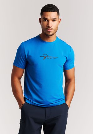 Man with short curly hair wearing a bright blue "Protest" logo T-shirt, standing with hands in dark pants pockets against a plain background.