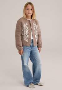 Fată blondă îmbrăcată într-o jachetă maro matlasată cu broderie albă, blugi largi, de culoare albastru deschis, cu tăieturi, și balerini cu mărgele, stând.