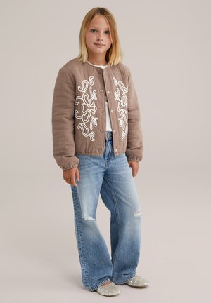 Blondes Mädchen trägt eine braune gesteppte Jacke mit weißer Stickerei, hellblaue, verzierte Schlagjeans und mit Pailletten besetzte Ballettschuhe und steht.
