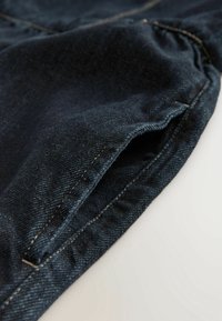 Gros plan sur un tissu en denim bleu foncé montrant une poche avant avec des coutures visibles sur un fond blanc.