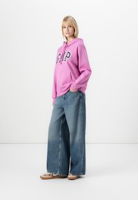 Różowa bawełniana bluza z kapturem z logo "GAP", z bocznymi kieszeniami, połączona z szerokimi niebieskimi dżinsami i beżowymi trampkami. Prosty design, bez wzorów.