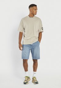 Beżowy bawełniany t-shirt z czarnym napisem na kołnierzyku, w komplecie z jasnoniebieskimi dżinsowymi szortami. Noszony z butami z mieszanych materiałów i białymi skarpetkami.