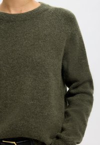 Personne portant un pull doux, duveteux, vert olive avec un col rond et des manches longues sur un fond clair uni.