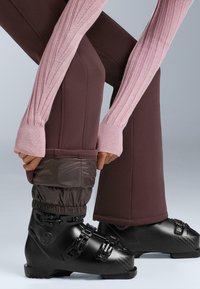 Scarponi da sci neri con più fibbie, abbinati a pantaloni marroni aderenti e un maglione a costine rosa, che mostrano un approccio allo stile a strati.