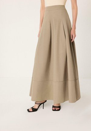 Femme portant une jupe longue beige taille haute plissée, associée à des sandales à talons hauts noires ouvertes aux orteils, debout devant un fond uni.