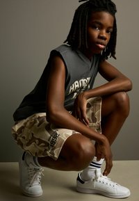 Grijze mouwloze shirt, camouflagebroek in beige tinten, witte sneakers met zwarte accenten, geposeerd door een hurkende persoon tegen een neutrale achtergrond.