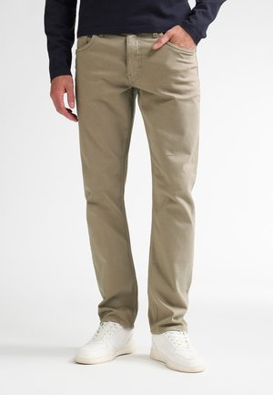 Hombre que lleva pantalones beige, zapatillas blancas y una camisa de manga larga oscura con una mano en el bolsillo, de pie sobre un fondo blanco.