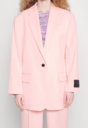 Person iført en pastelrosa oversized blazer med en enkelt sort knap og matchende bukser samt en flerfarvet striped top under.