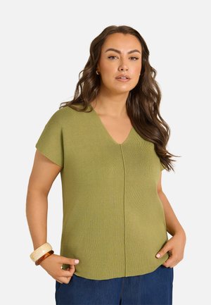Femme aux longs cheveux ondulés portant un haut vert olive à manches courtes et col en V ainsi qu'un pantalon bleu, posant avec les mains près des poches et des bracelets au poignet.