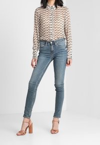 Camicia trasparente beige con bottoni e motivo geometrico, abbinata a jeans slim-fit blu chiaro e sandali con tacco marroni.