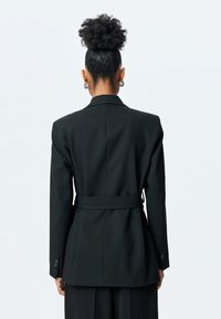 Chaqueta negra ajustada con cintura ceñida, solapas muesca y mangas largas, que exhibe una textura suave y un diseño estructurado.