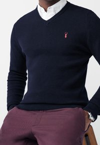 Laivastonsininen neulepaita V-kaula-aukkolla, teksturoidulla kankaalla ja pienellä punaisella hummarilogolla rinnassa, yhdistetty viininpunaisiin housuihin.