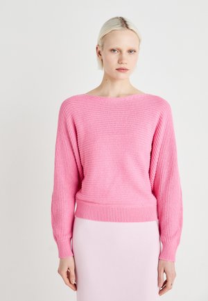 Pink knitted sweater with a loose fit, textured design, ribbed cuffs and hem, featuring a wide neckline. Worn over a pink skirt.