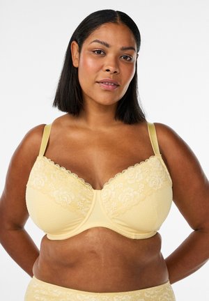 Ženska, oblečena v svetlo rumen čipkast modrček in usklajena spodnja perila, stoji z rokami na bokih ob belem ozadju.