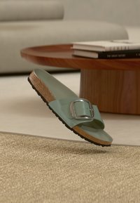 Πράσινο γυαλιστερό σανδάλι χωρίς λουριά με μεταλλική αγκράφα, πάτο από φελλό, μαύρη ελαστική σόλα και λεία υφή, αιωρούμενο πάνω από πλεκτή επιφάνεια.