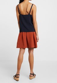 Námořnické modré a rezavě zbarvené bezrukávové šaty s rozšířenou sukní. Obsahují nastavitelné ramínka a strukturovaný vršek, hladkou tkaninu na spodní části.