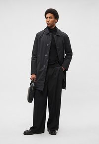 Fekete vízálló kabát gombokkal, fekete garbóval és bőszárú nadrággal párosítva. Fekete aktatáska a kézben; texturált cipők.