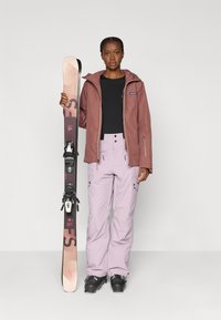 Skiausrustning: en rustfarvet vandtæt jakke, sort top, lyse lilla bukser med lommer og sorte skistøvler. Personen holder ski med bindinger.