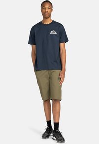 Marinblå bomulls t-shirt med vitt tryck på bröstet, i kombination med olivgröna shorts. Modellen bär svarta sneakers och knähöga strumpor.