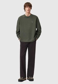 Felpa verde oliva in tessuto morbido, con scollo tondo e dettaglio grafico sul petto. Abbinata a pantaloni neri e scarpe con motivo.