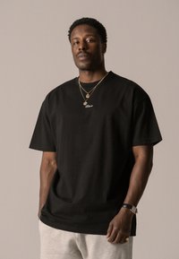 Black cotton t-shirt with short sleeves and a crew neck, featuring a small white logo. Gray sweatpants and gold jewelry accent the look.