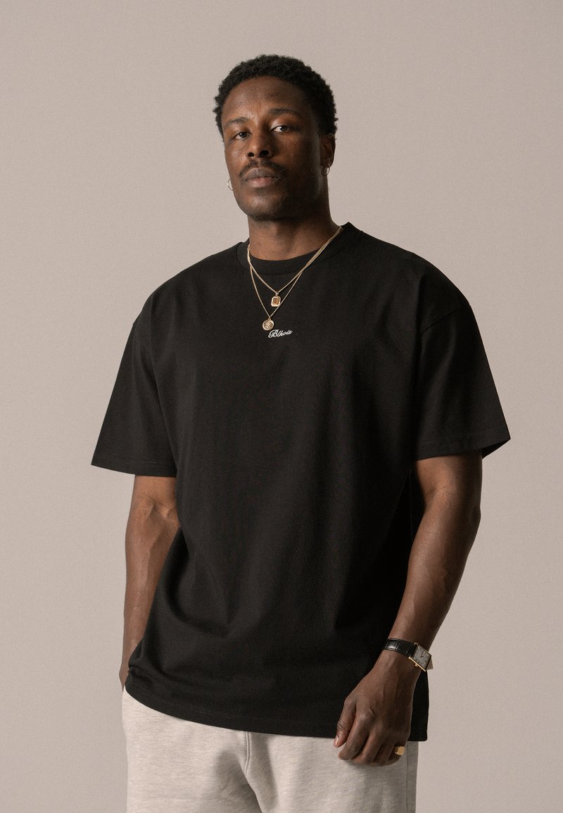 Black cotton t-shirt with short sleeves and a crew neck, featuring a small white logo. Gray sweatpants and gold jewelry accent the look.