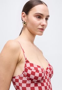 Γυναίκα με μαλλιά πίσω χτενισμένα, που φοράει μεγάλα χρυσά σκουλαρίκια σε σχήμα αστεριού και κόκκινο-ροζ καρό αμάνικο τοπ.